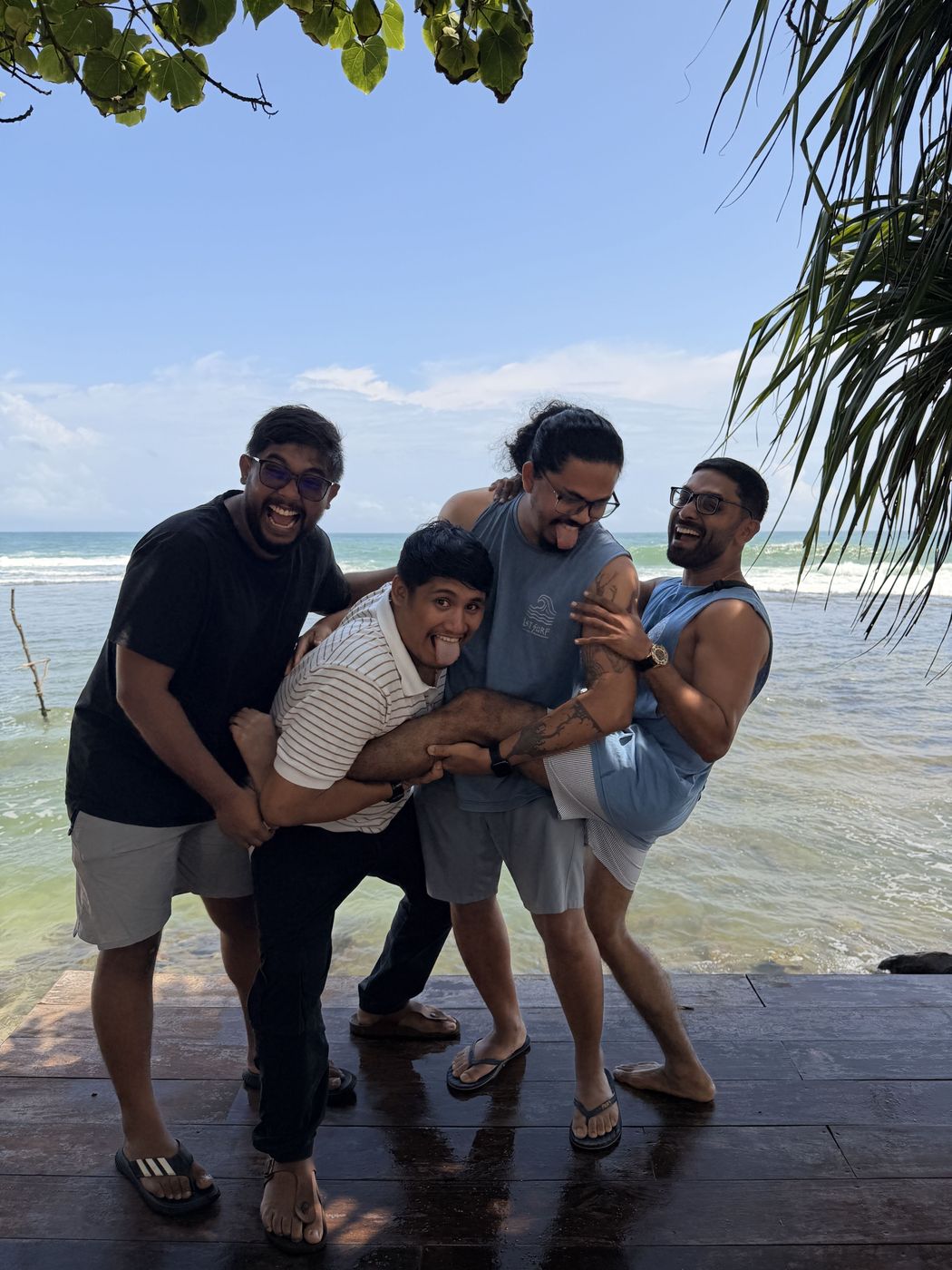Four team members laughing and mucking about together on a beach deck under palm fronds, the ocean behind them.