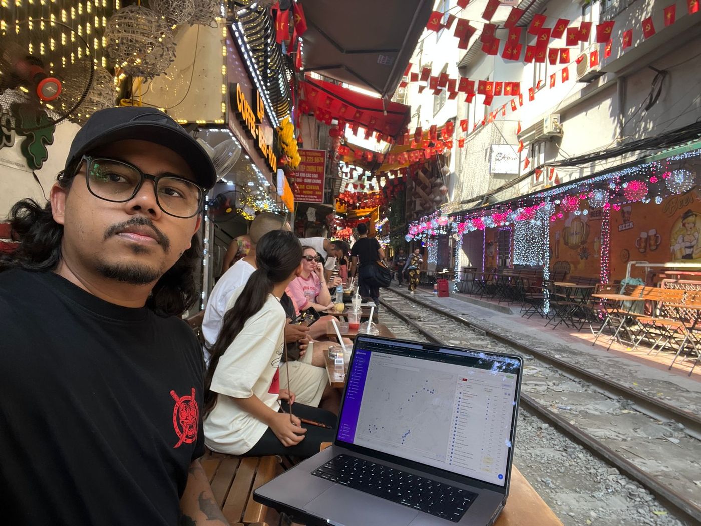 A team member in a cap taking a selfie at an outdoor table beside a railway track lined with red lanterns and market stalls, a laptop open in front of him.