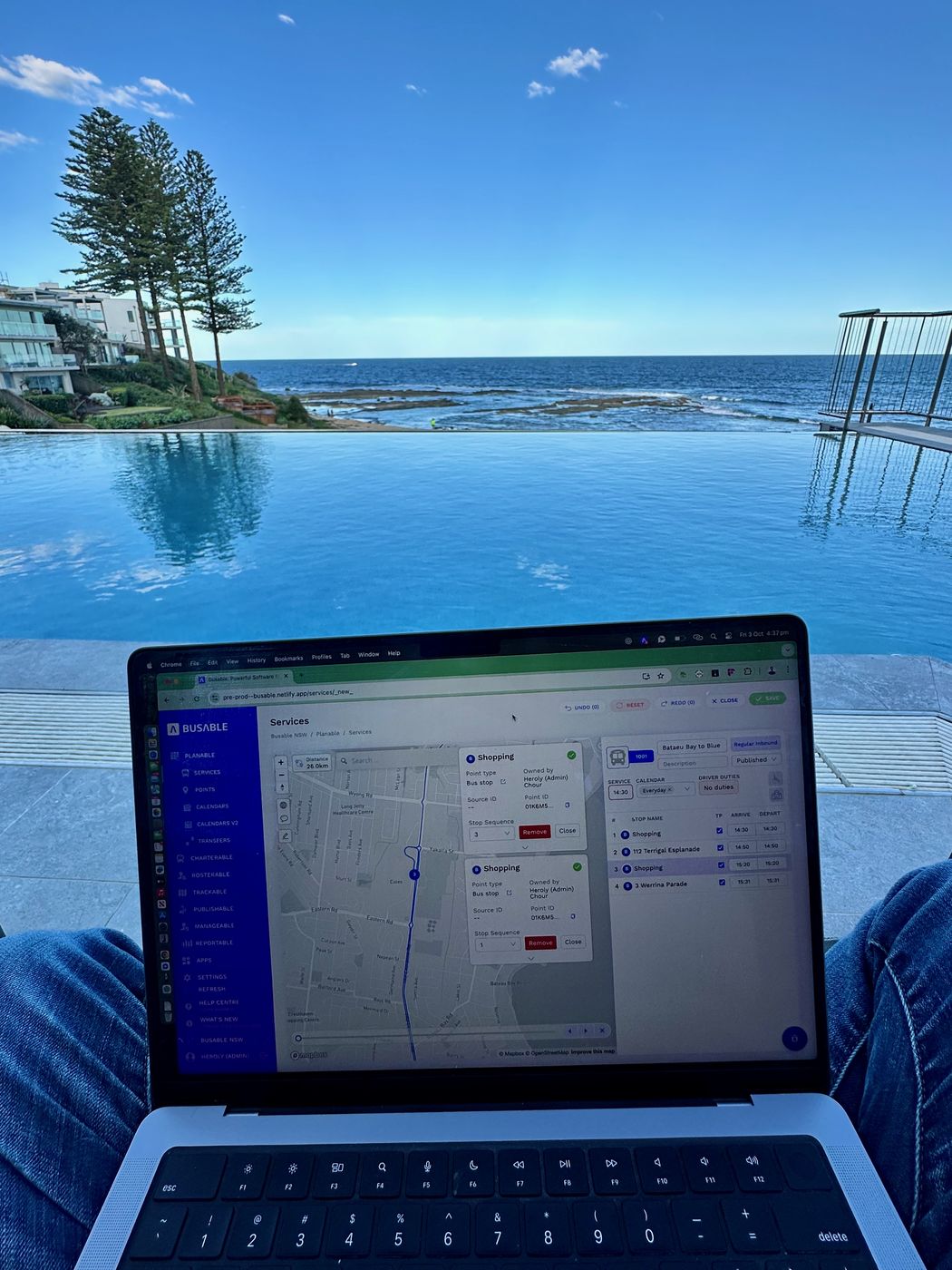 A laptop resting on someone’s lap showing the Busable services screen with a route map, an ocean pool and the sea behind it.
