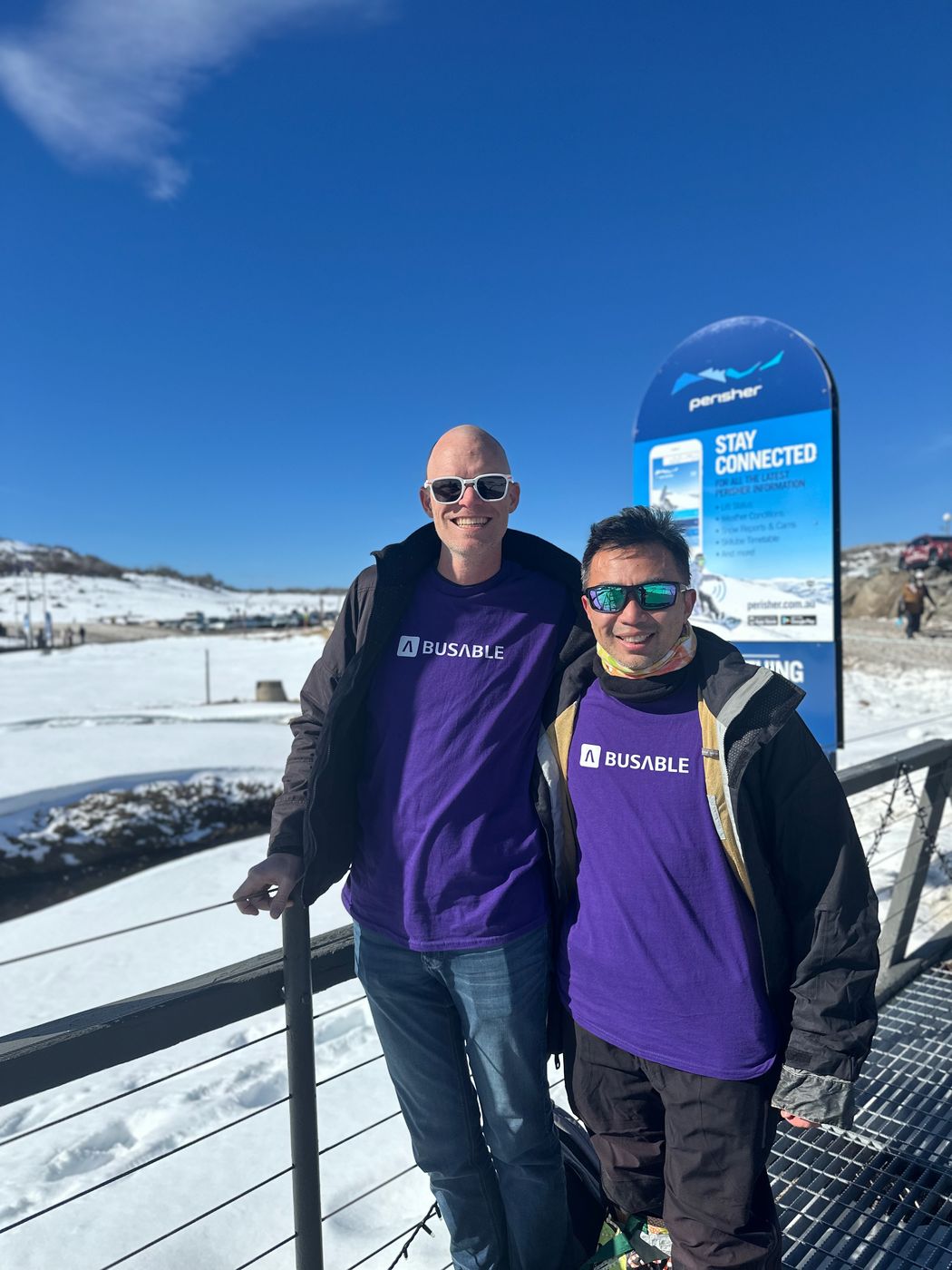 Two team members in purple Busable t-shirts standing on a snow-covered slope at Perisher under a clear blue sky, beside a Perisher sign reading Stay Connected.