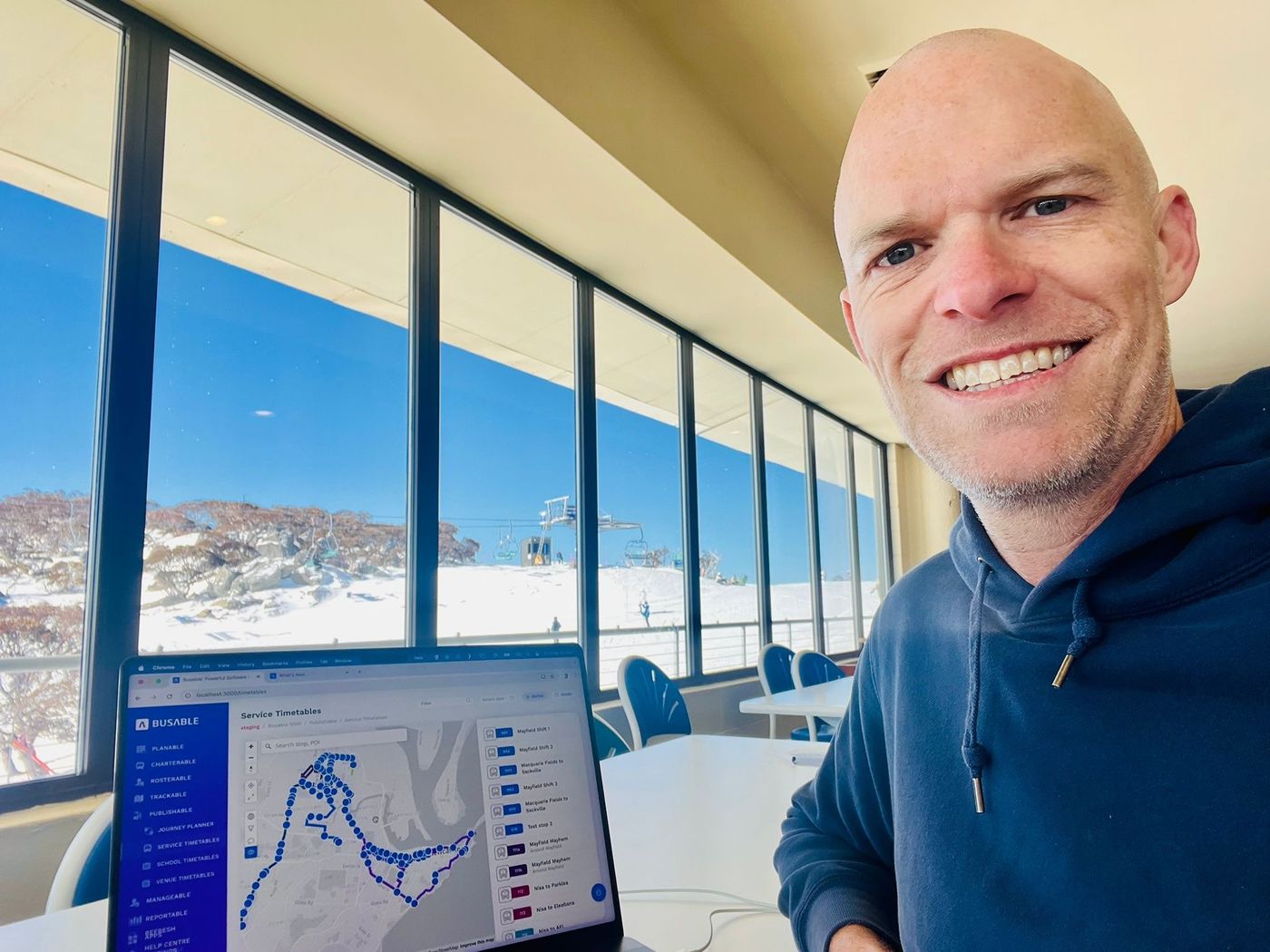 A team member smiling at the camera in a lodge with floor-to-ceiling windows looking out over a snowfield, a laptop on the table showing the Busable service timetables screen.
